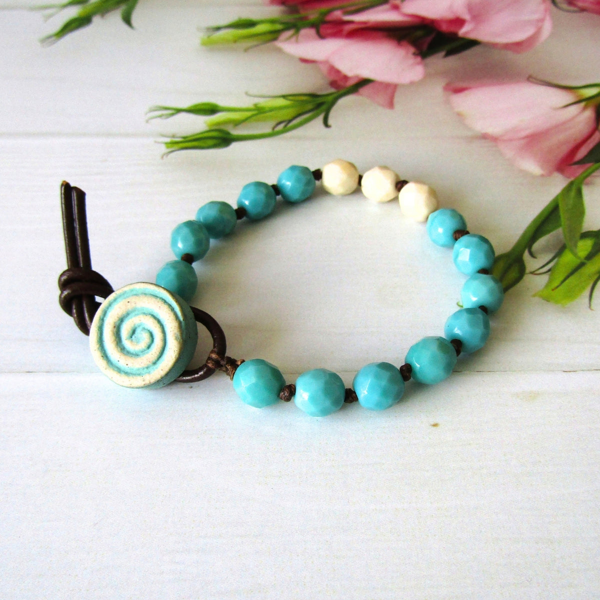 A beaded bracelet with turquoise blue faceted beads, a spiral-patterned turquoise and cream button, and dark brown cord, displayed on a white wooden surface with pink flowers in the background.