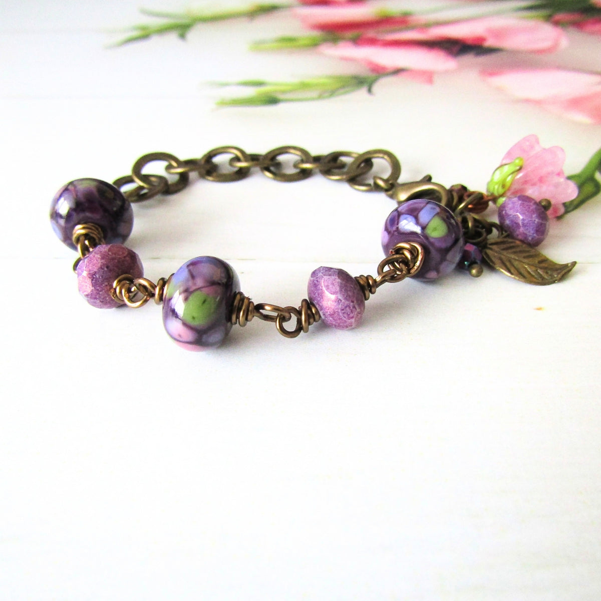 A handmade beaded chain bracelet with bronze-toned metal links, featuring purple floral glass beads, faceted purple beads, and a small leaf charm, displayed on a white surface with blurred pink flowers in the background.