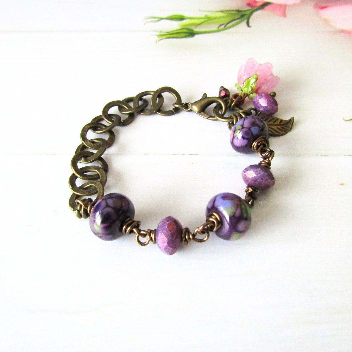 An antique bronze chain bracelet with purple artisan made beads and a small pink flower charm, displayed on a white wooden surface with soft pink flowers in the background.