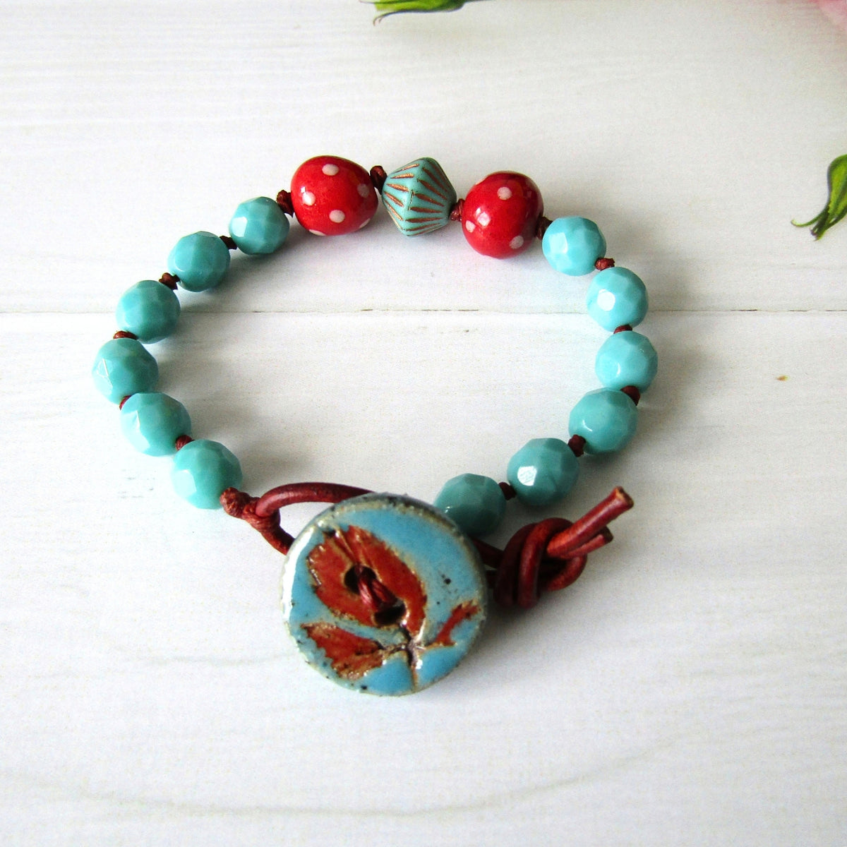 A handmade beaded bracelet with turquoise and red beads, featuring a large blue & red button clasp, displayed on a white wooden surface.