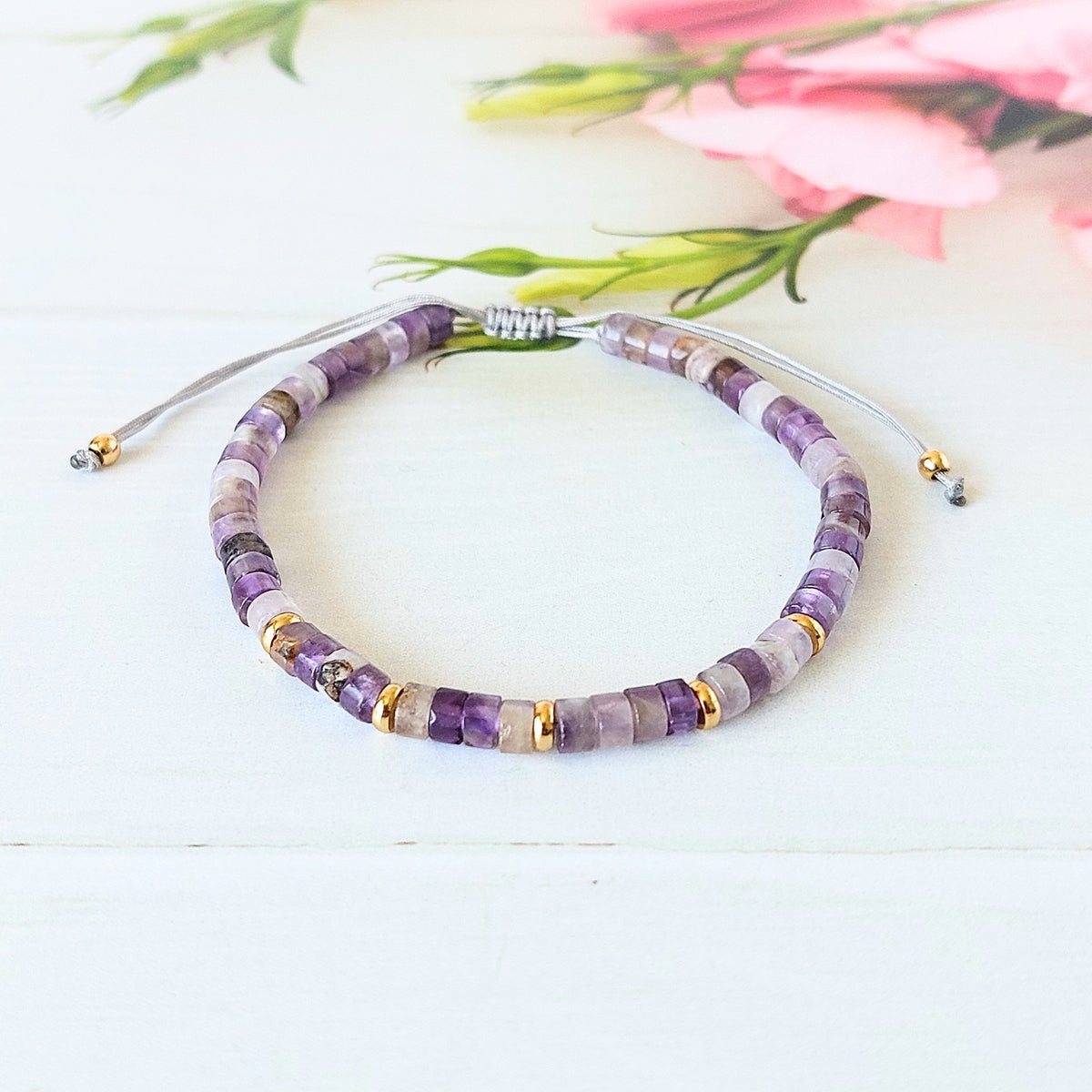 A beaded cord bracelet made of purple and lavender amethyst gemstone beads with small gold spacer rings, displayed on a white wooden surface with soft pink flowers in the background.