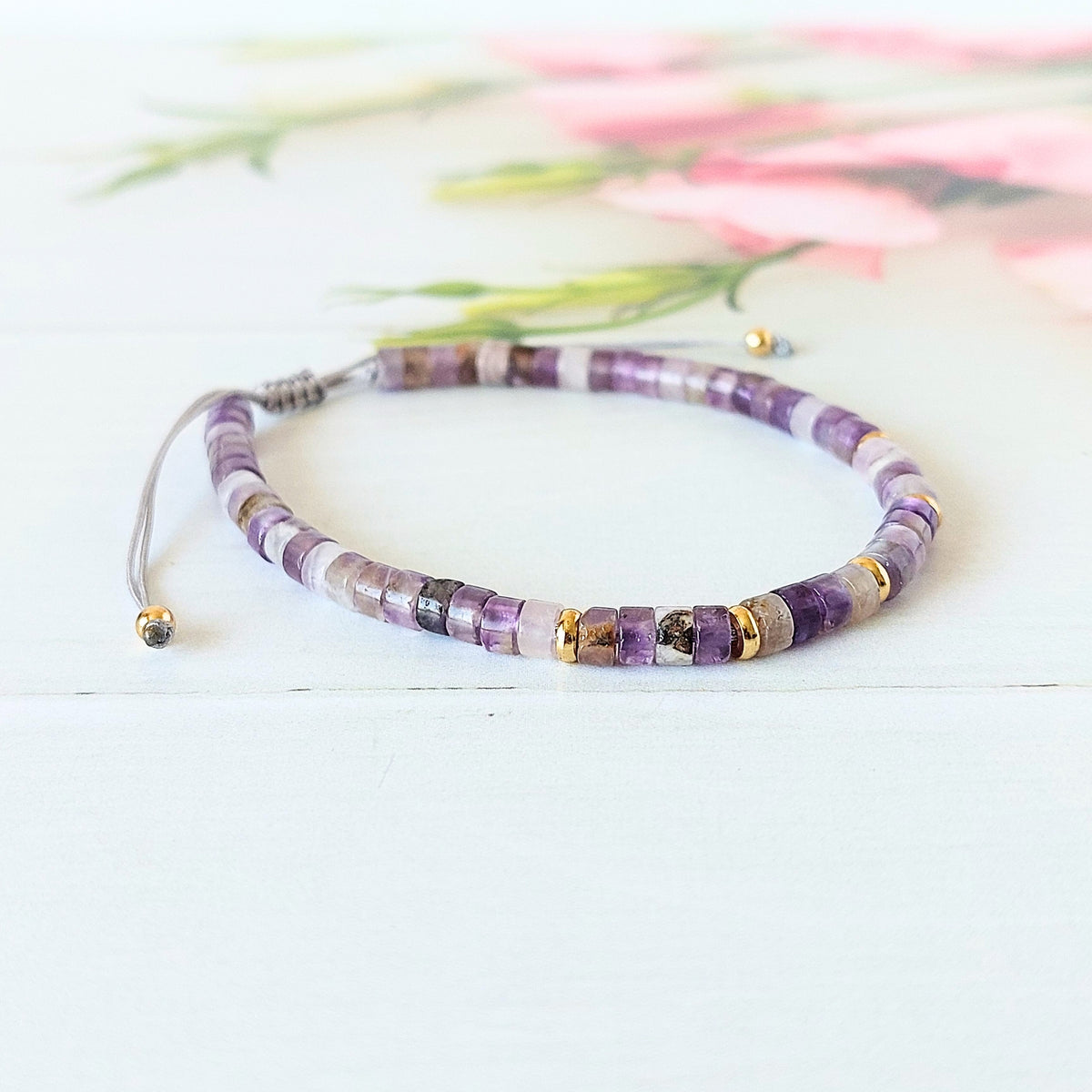 A beaded gemstone bracelet made of purple, white, and grey amethyst beads with small gold spacer accents, displayed on a white wooden surface with a soft pink floral background.