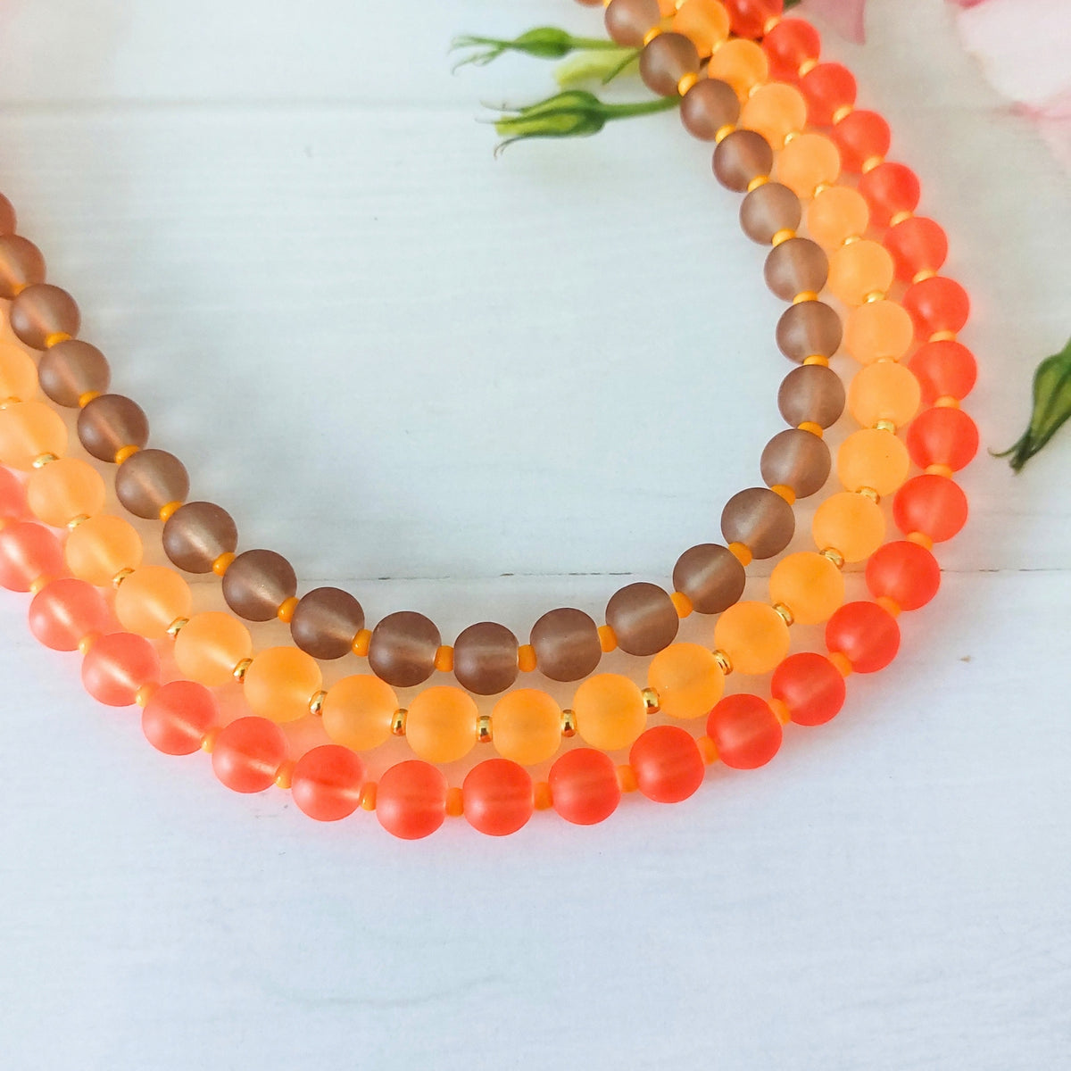 Three round beaded necklaces arranged in a circle on a white wooden surface. The necklaces feature warm brown beads, soft amber glass beads and lively coral-tone beads.