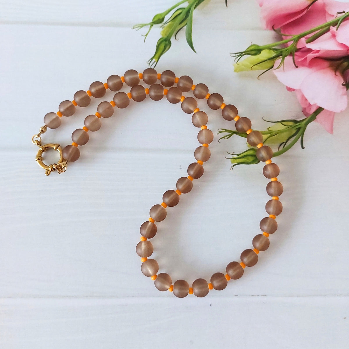A beaded necklace made of frosted brown glass beads separated by small orange spacer beads, with a gold clasp, arranged on a white wooden surface next to pink flowers.