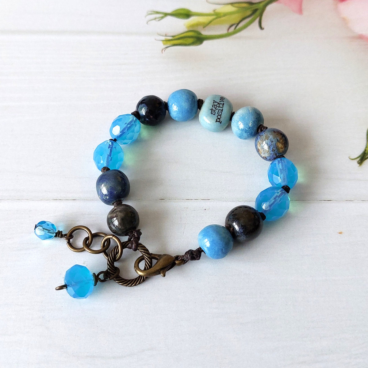 A beaded bracelet with light blue, dark blue, and dark grey beads, featuring a antique brass clasp and two dangling blue beads, displayed on a white wooden surface.