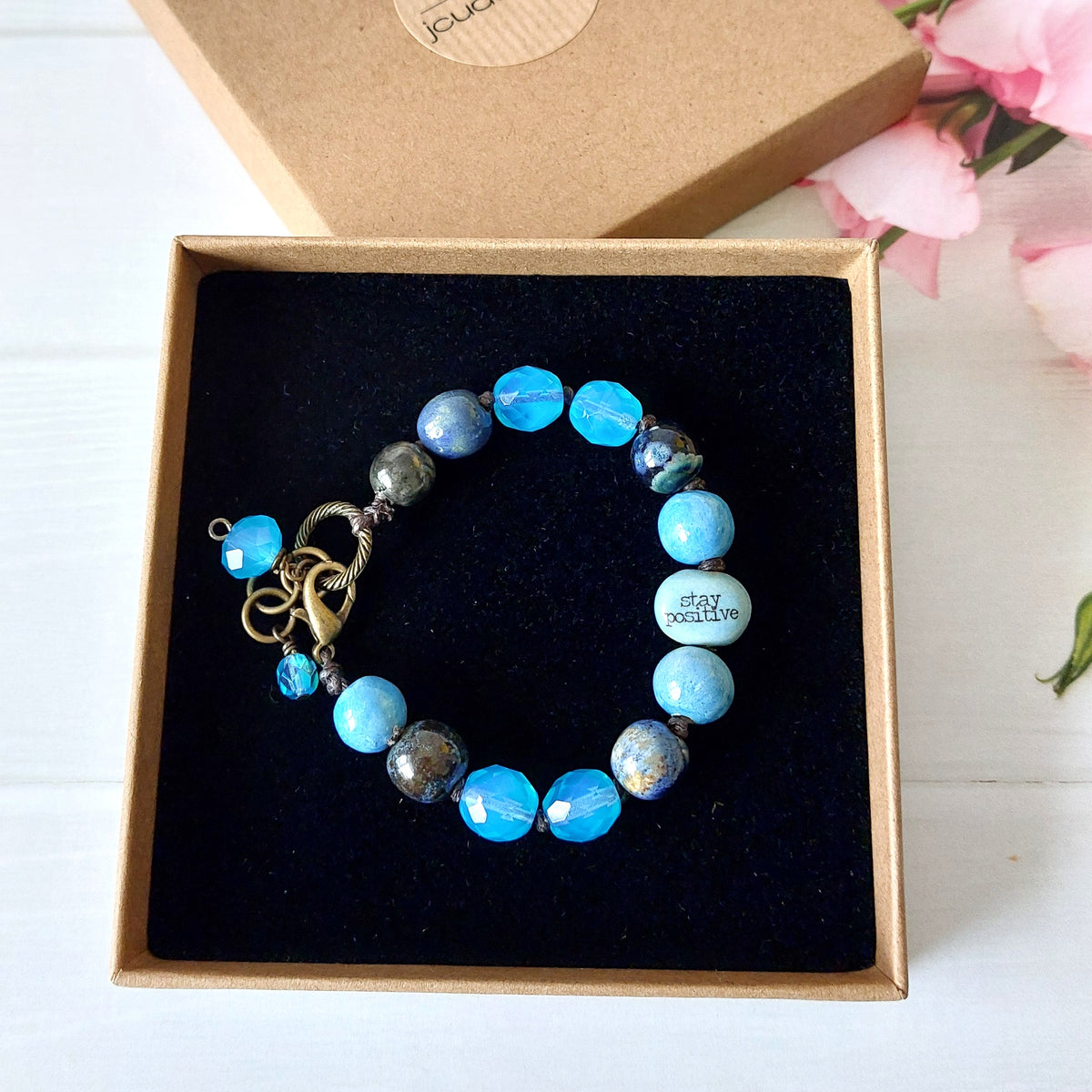 A beaded bracelet with light blue, dark blue, and grey beads, displayed inside a black-lined kraft gift box. The bracelet features an antique brass clasp and a small dangling blue bead.