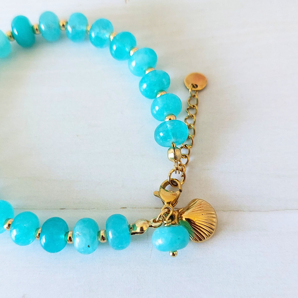 A turquoise beaded bracelet with gold-plated stainless steel spacer beads, a gold lobster clasp, and a gold shell charm, displayed on a white wooden background.
