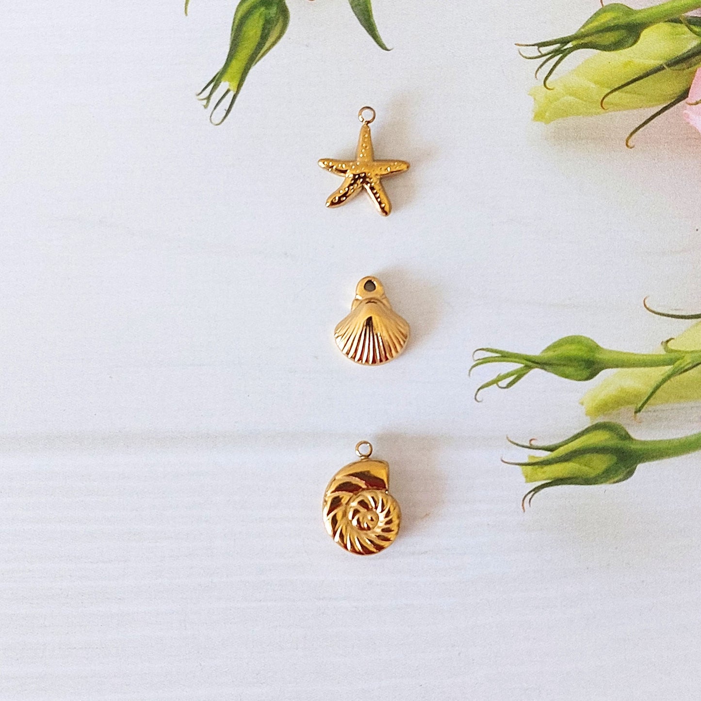 Three small gold-toned jewelry charms shaped like a starfish, a scallop shell, and a spiral shell, arranged vertically on a white background with green flower buds as decorative props.