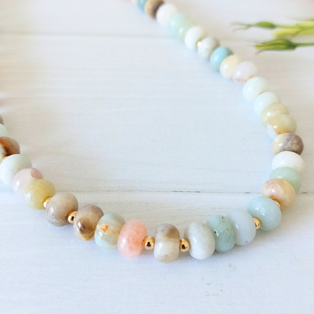 A beaded necklace made of polished pastel gemstone beads in white, blue, peach, and taupe, separated by small gold-plated stainless steel spacer beads, displayed on a white wooden surface.