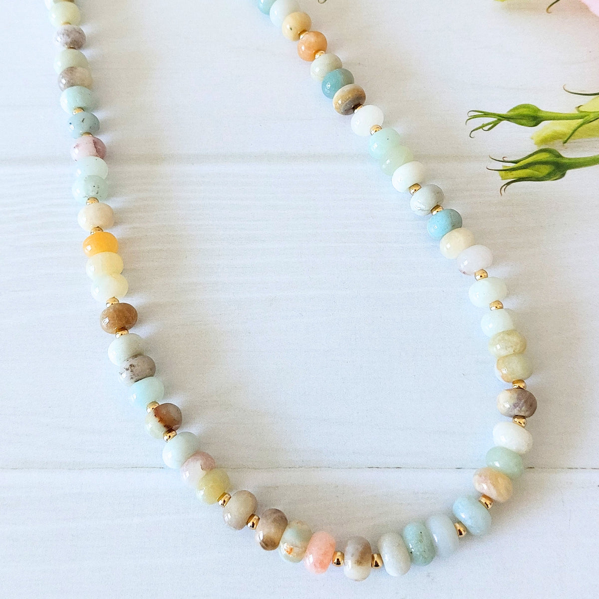 A beaded necklace made of polished rondelle gemstone beads in pastel shades of mint, peach, blue, cream, and brown, separated by small gold-plated stainless steel spacer beads, displayed on a white wooden surface.