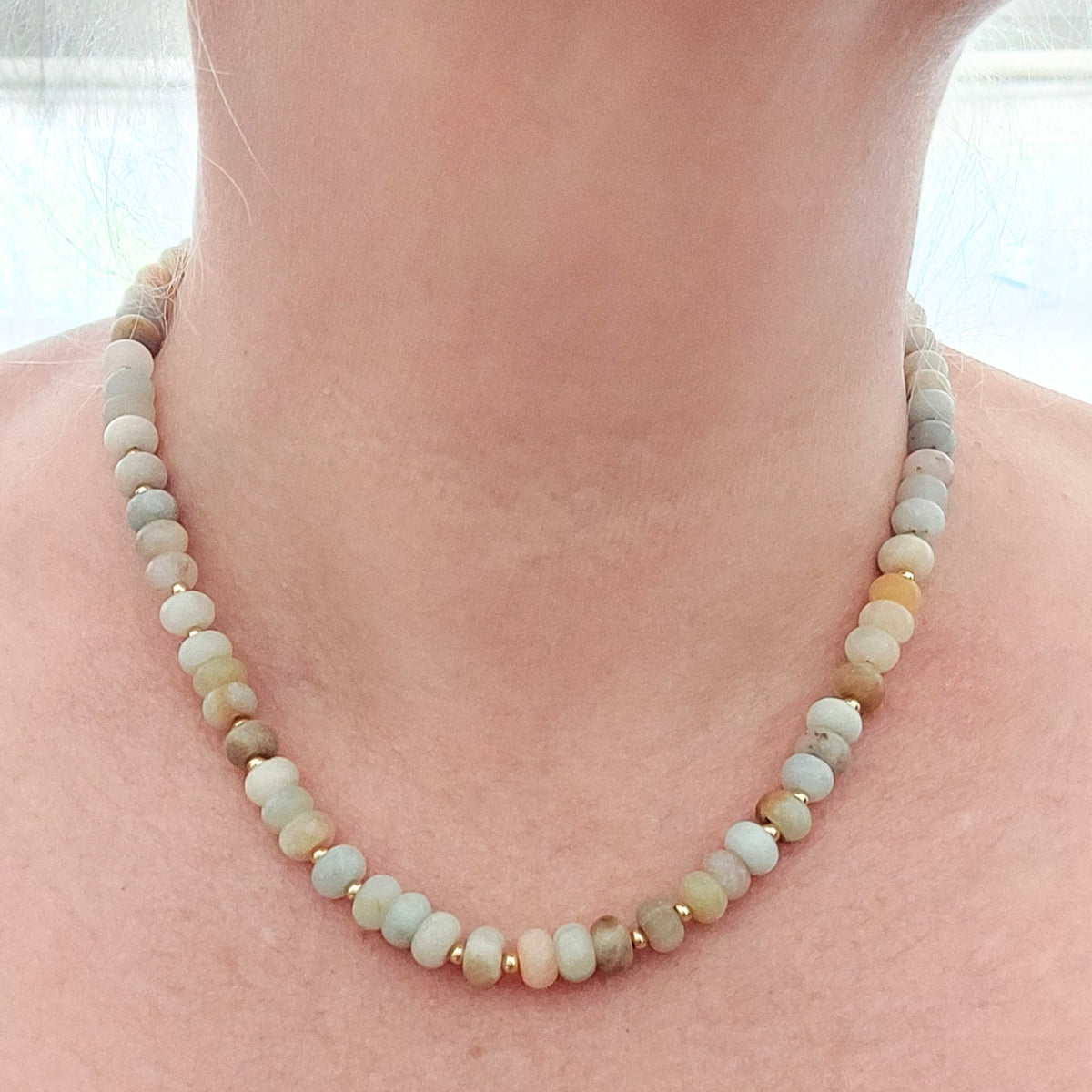 A close-up of a person's neck and collarbone wearing a beaded necklace made of small rondelle gemstone beads in muted sage green, cream, tan, and light gray, separated by small gold-plated stainless steel spacer beads.