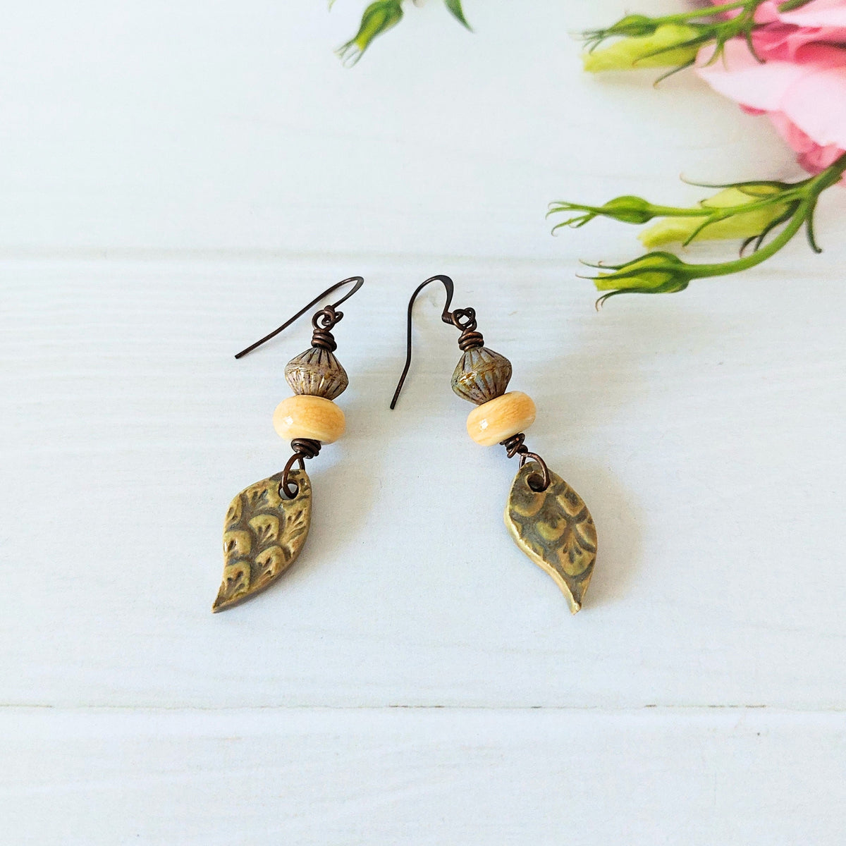 A pair of artisan drop earrings with olive ceramic tear-drop charms, light yellow lampwork glass beads, and dark olive bicone beads, on a white wooden surface.