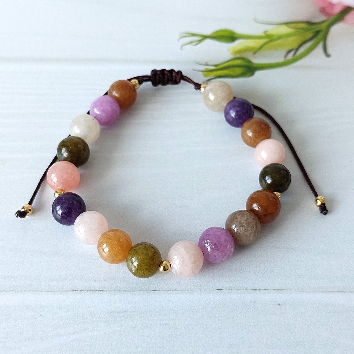 A multicolor beaded bracelet made of polished round gemstone beads in pink, purple, brown, green, white, and orange, with gold spacer beads and a dark brown adjustable cord, displayed on a light blue-white wooden surface with a small green flower in the background.
