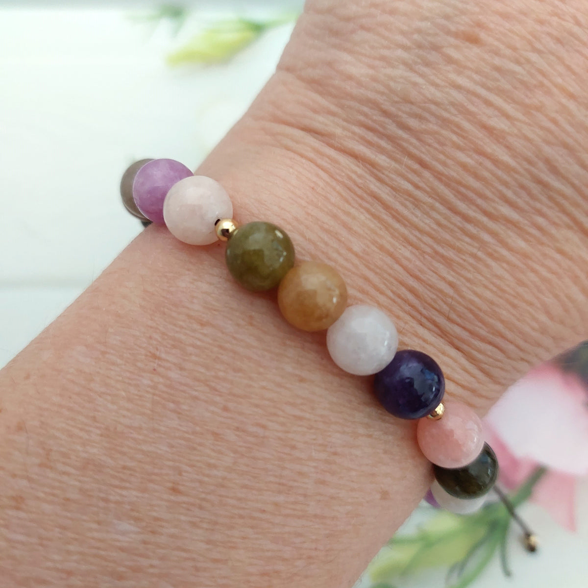 A close-up of a beaded gemstone bracelet worn on a person's wrist. The bracelet features smooth, polished round beads in purple, pale pink, olive green, tan, white, and dark blue, separated by small gold spacer beads.