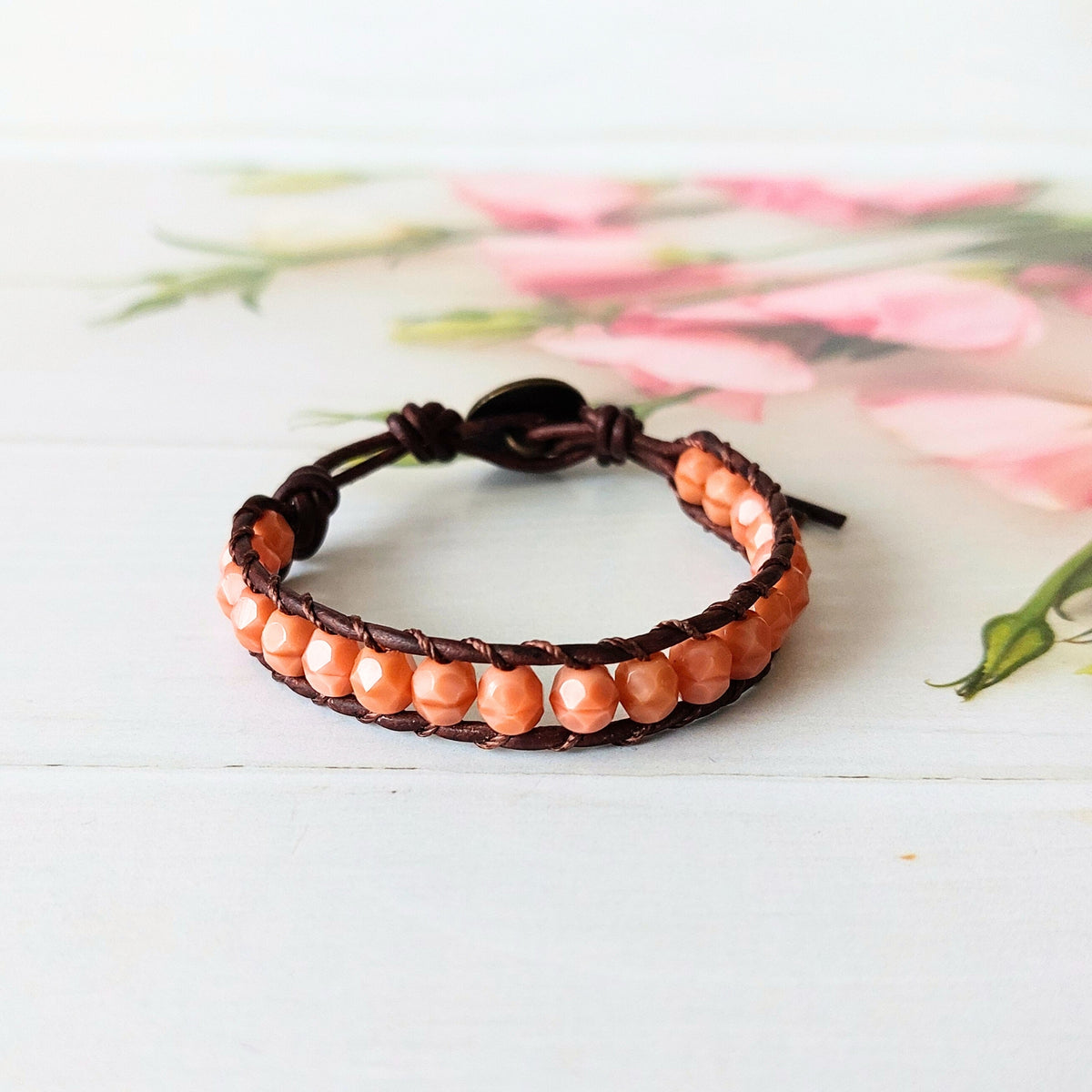 A handmade beaded wrap bracelet with dark brown leather cords, soft coral beads, and a decorative brass metal button fastener, displayed on a white wooden surface with a soft pink floral background.