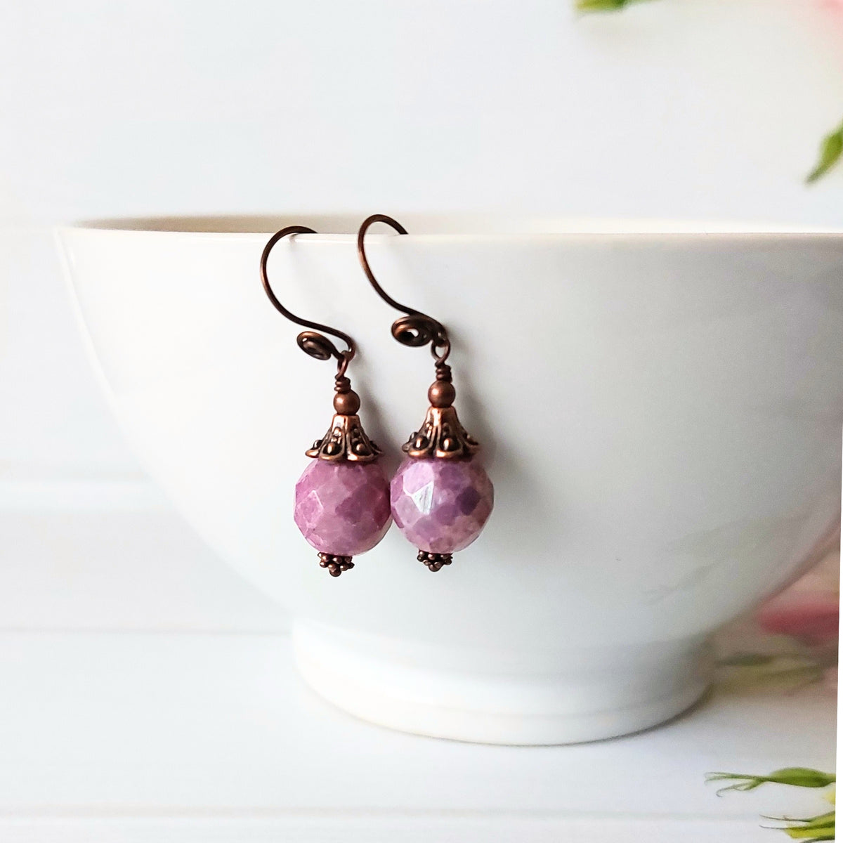 A pair of dangling earrings with faceted light purple beads and copper scrolled earwires, hanging from the rim of a white ceramic bowl on a white surface.
