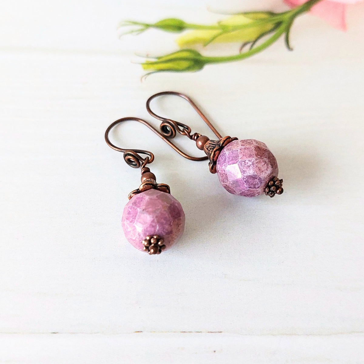 A pair of handmade dangle earrings featuring faceted purple beads and copper-toned wire hooks, displayed on a white wooden surface with soft green flower buds in the background.