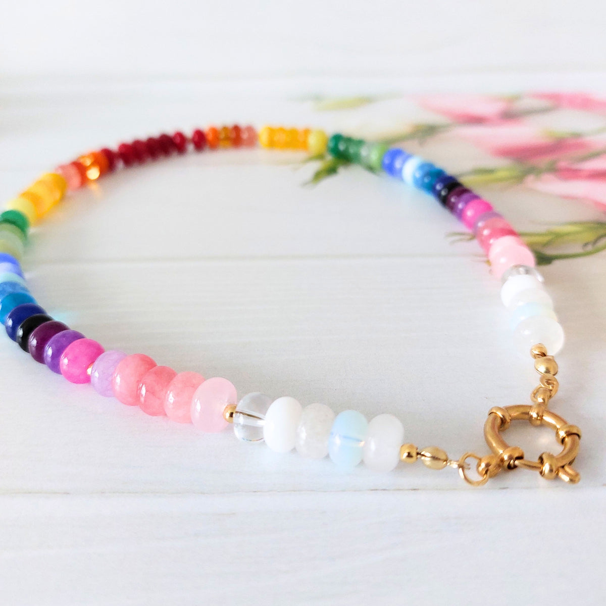 A beaded necklace with rainbow-colored round beads, a cluster of white and light blue beads, and a gold toggle clasp, displayed on a white wooden surface.