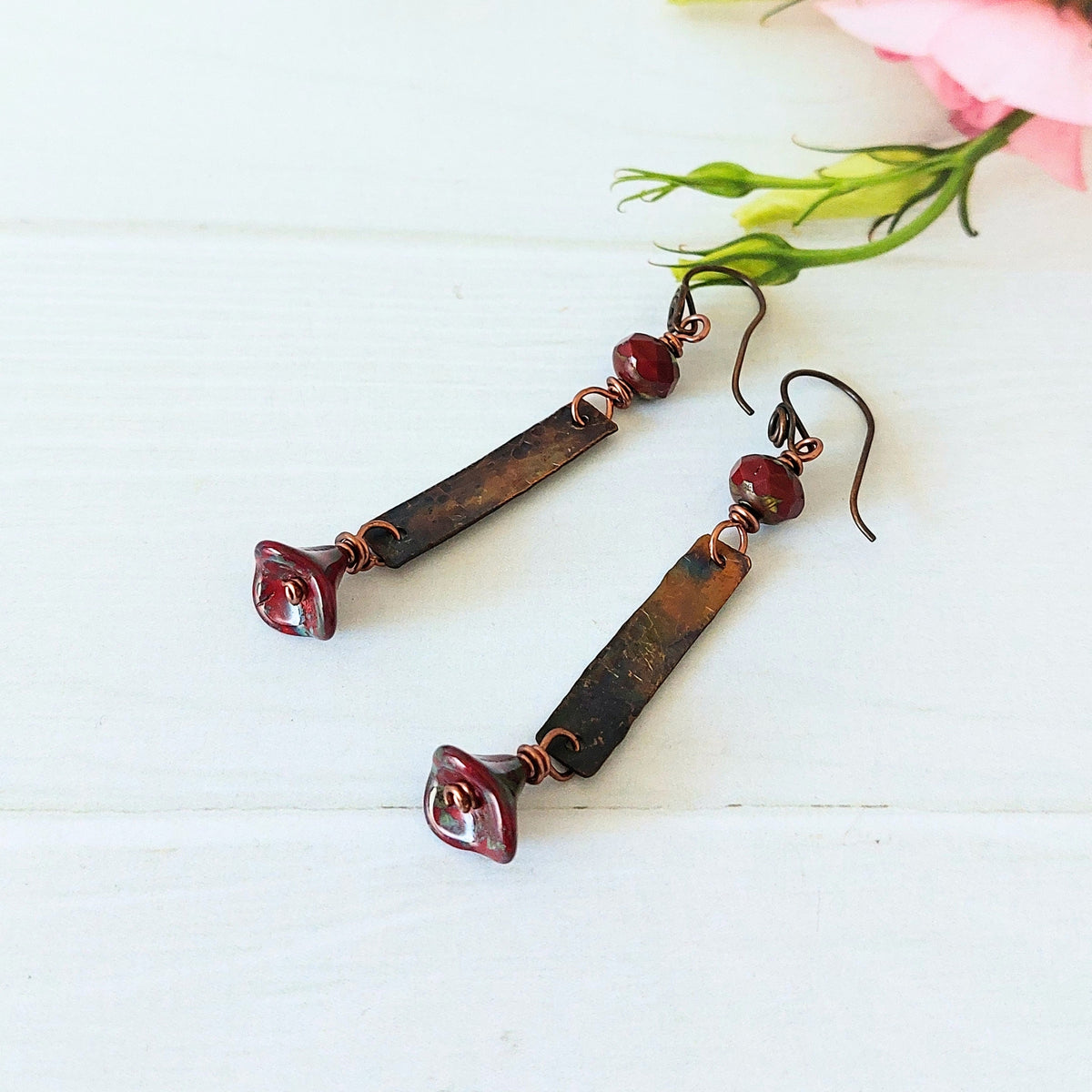 A pair of handmade dangle earrings with dark brown rectangular bars, copper wire accents, small faceted red beads, and flower marbled red glass beads, displayed on a white wooden surface with a pink flower in the corner.
