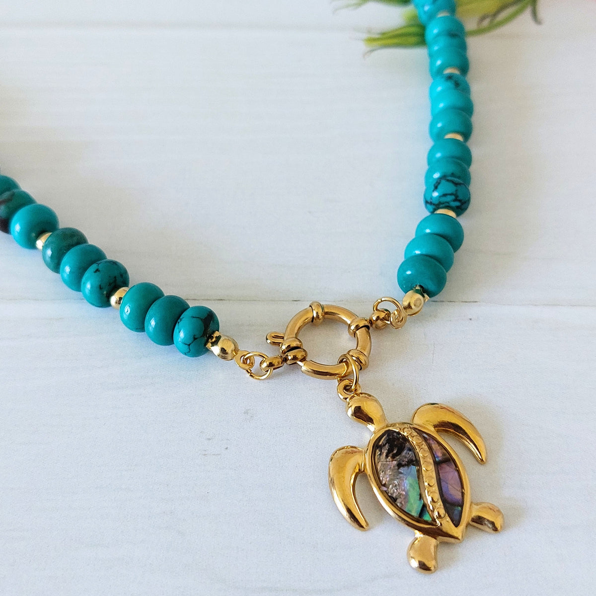 Close up of turquoise howlite bead necklace with a gold turtle pendant featuring an iridescent shell inlay, displayed on a white wooden surface.