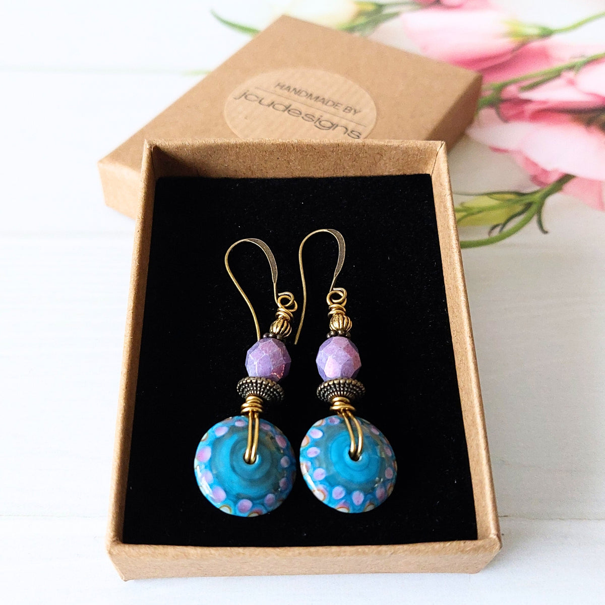 A pair of handmade dangle earrings displayed in an open kraft gift box. The earrings feature gold-tone fishhook wires, small lavender beads, and round turquoise blue glass disc beads with purple dots.