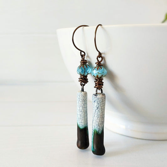 A pair of handmade dangle earrings with turquoise blue faceted beads, brass wire wrapping, and crackled-effect ceramic drops that fade from pale blue to bronze and black, hanging from a white ceramic bowl.