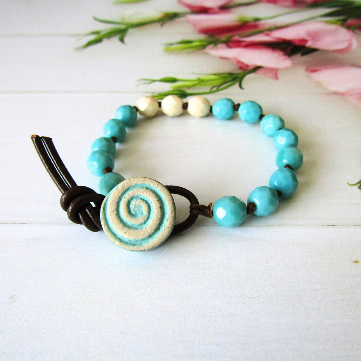 A handmade beaded bracelet with turquoise blue & cream beads, a central round beige ceramic button with a blue swirl design, and a dark brown leather loop closure, displayed on a white wooden surface with pink flowers in the background.