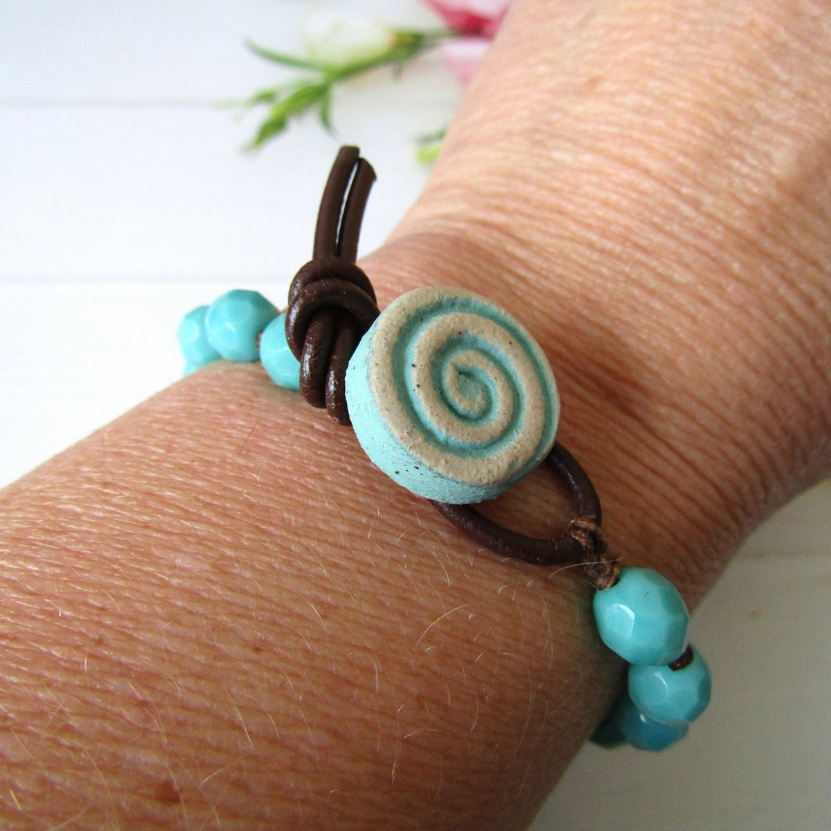 A close-up of a person's wrist wearing a handmade bracelet with turquoise beads, a dark brown leather cord, and a round light blue and cream spiral button clasp.