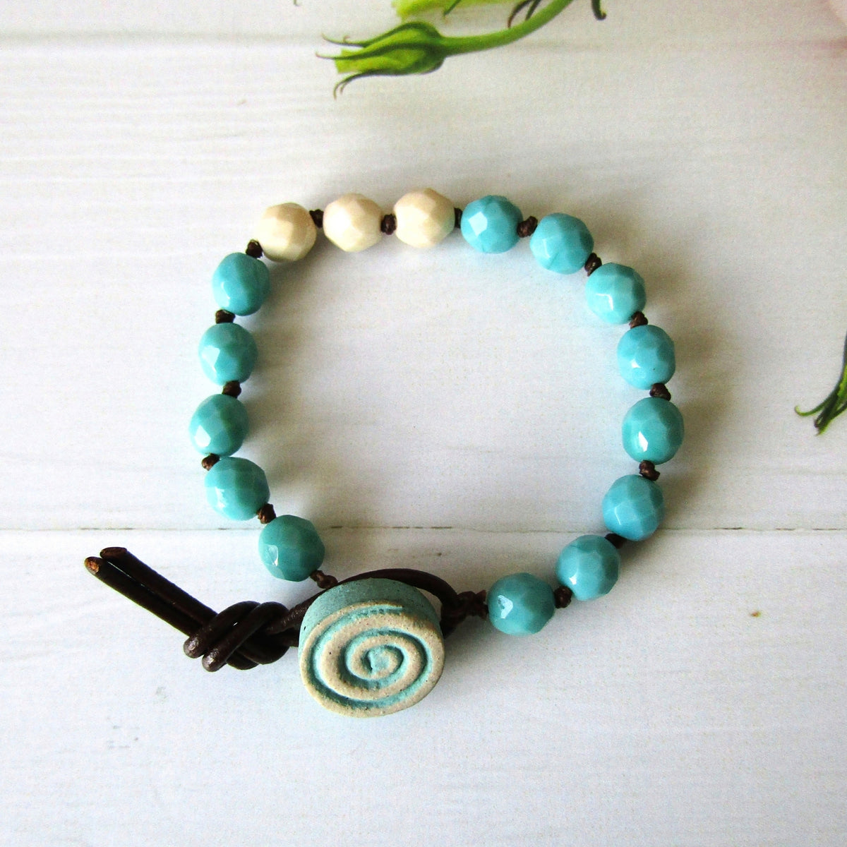 A handmade beaded bracelet with turquoise blue faceted beads, three cream-colored beads, a spiral-shaped turquoise button clasp, and a dark brown leather loop closure, displayed on a white wooden surface.