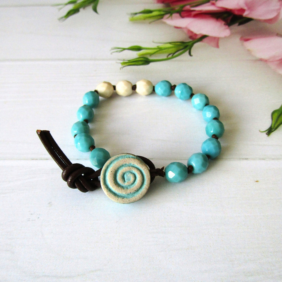 A beaded bracelet with turquoise blue faceted beads, a central round beige ceramic button with a blue spiral, and a dark brown cord closure, photographed on a white wood surface with pink flowers in the background.