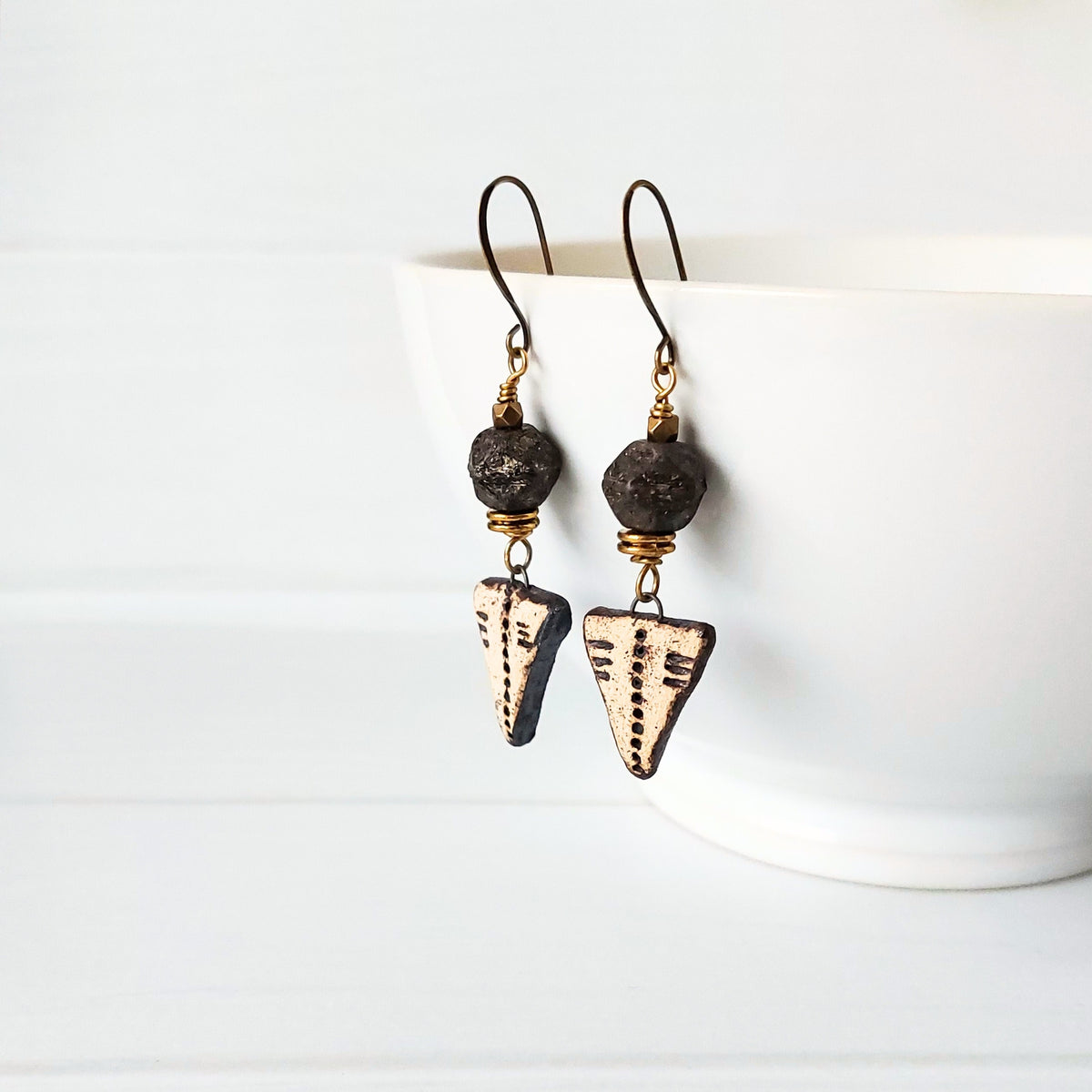 A pair of handmade dangle earrings with dark textured facted beads, brass wire spacers, and triangular rustic ceramic charms, displayed on a white ceramic bowl against a white background.