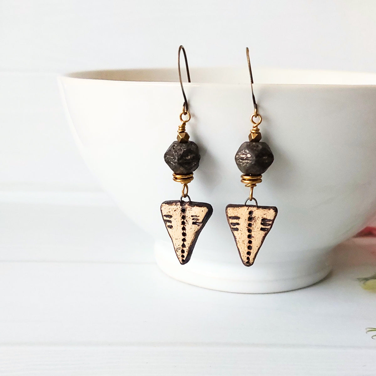 A pair of handmade dangle earrings with dark textured beads and triangular arrowhead charms featuring engraved details, displayed on a white ceramic bowl.