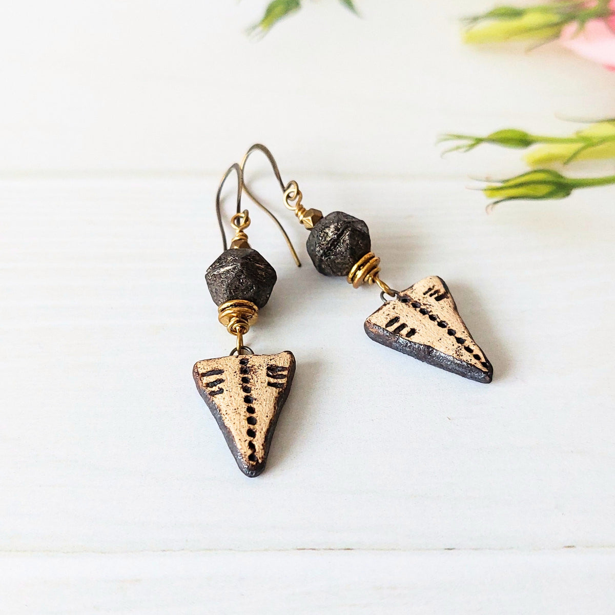A pair of handmade dangle earrings featuring triangular arrowhead charms with black speckled detailing on a distressed bronze finish, suspended from dark faceted beads and antique brass wire on fishhook wires, displayed on a white wooden surface with soft pink flowers in the background.