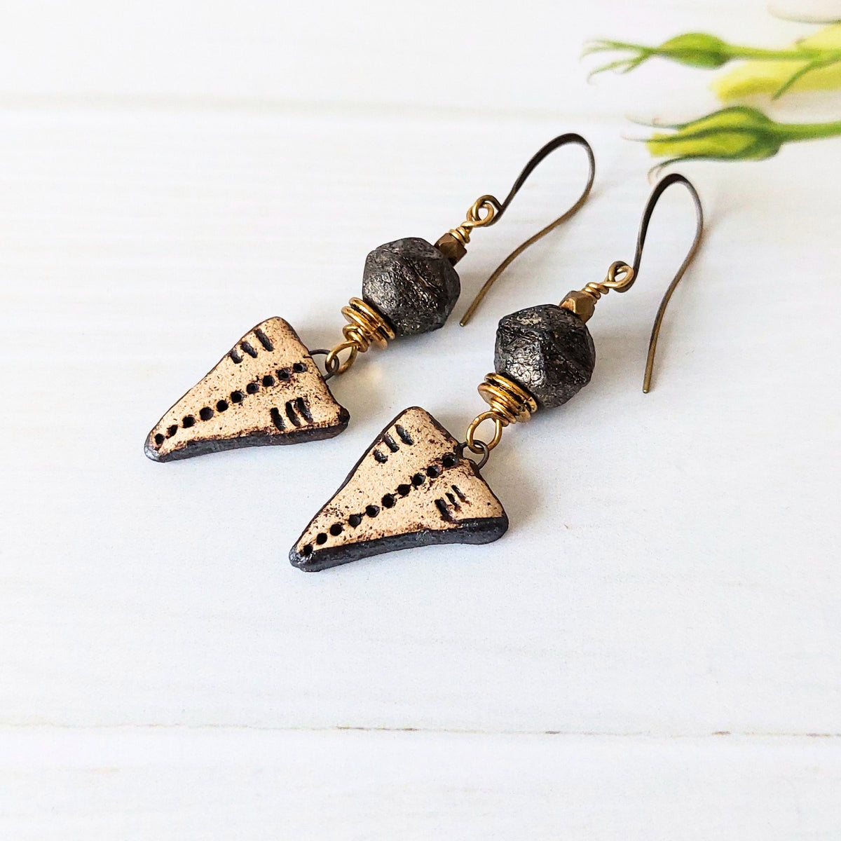A pair of handmade dangle earrings featuring textured dark metal beads and triangular arrowhead charms with stamped details, displayed on a white wooden surface.