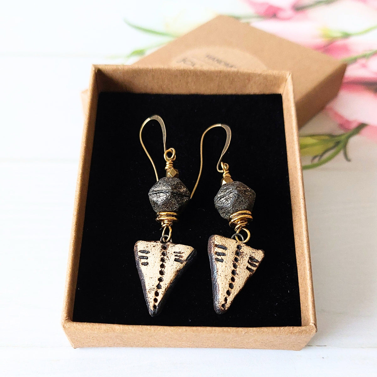 A pair of handmade dangle earrings displayed inside a brown kraft gift box. The earrings feature textured black beads, gold wire wrapping, and triangular cream ceramic drops with black engraved markings.