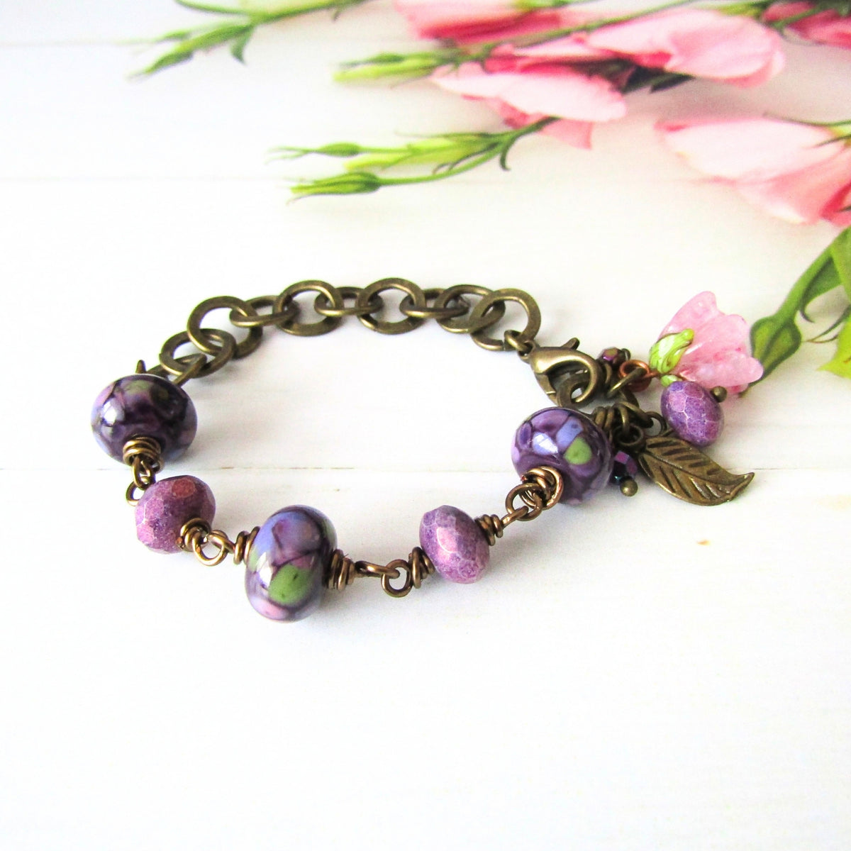 An antique bronze chain bracelet with purple floral lamp-work beads and a small leaf charm, styled on a white surface with pink flowers in the background.