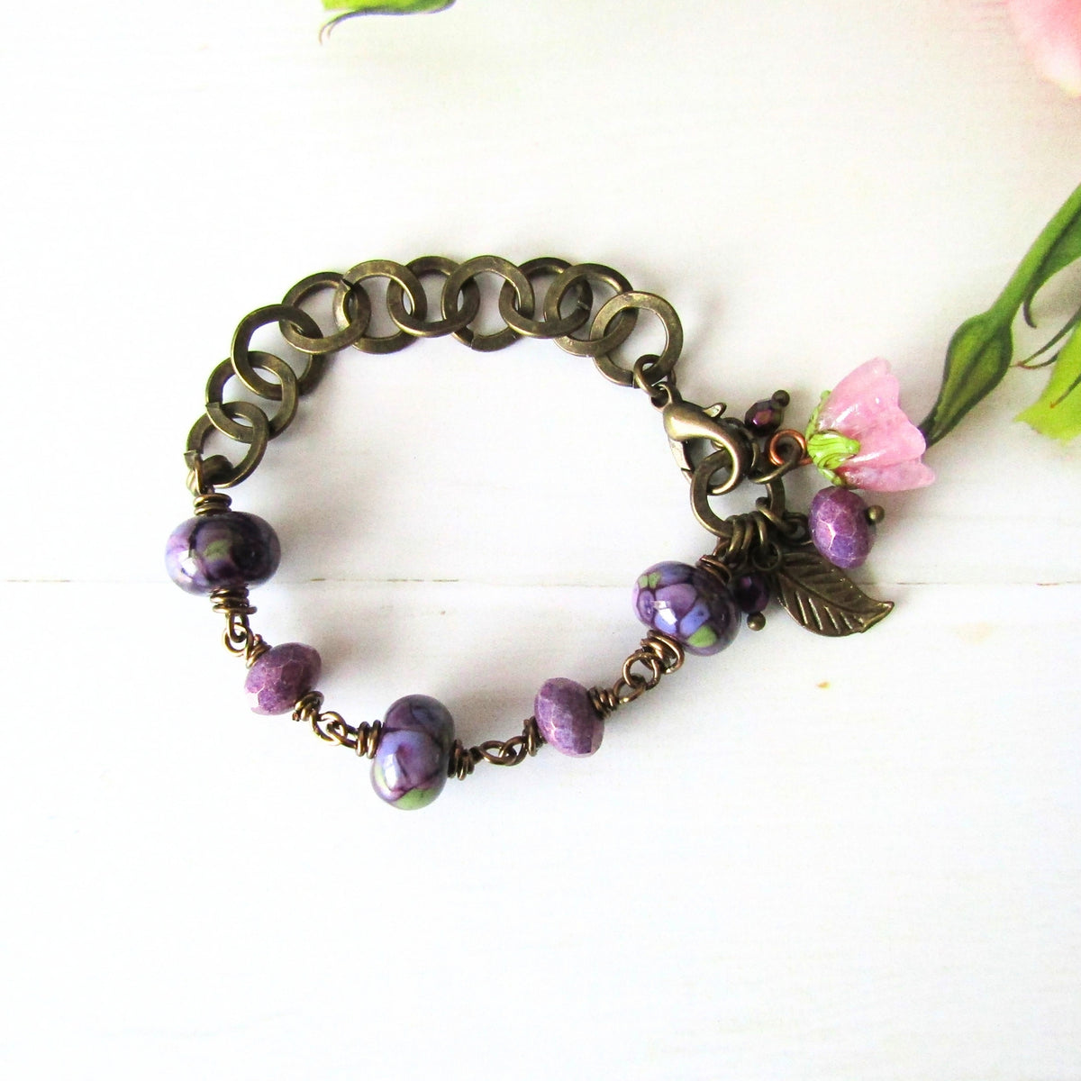 An antique bronze link bracelet featuring purple beads, a lobster clasp, and a dangling leaf charm with a pink flower accent, displayed on a white wooden surface.