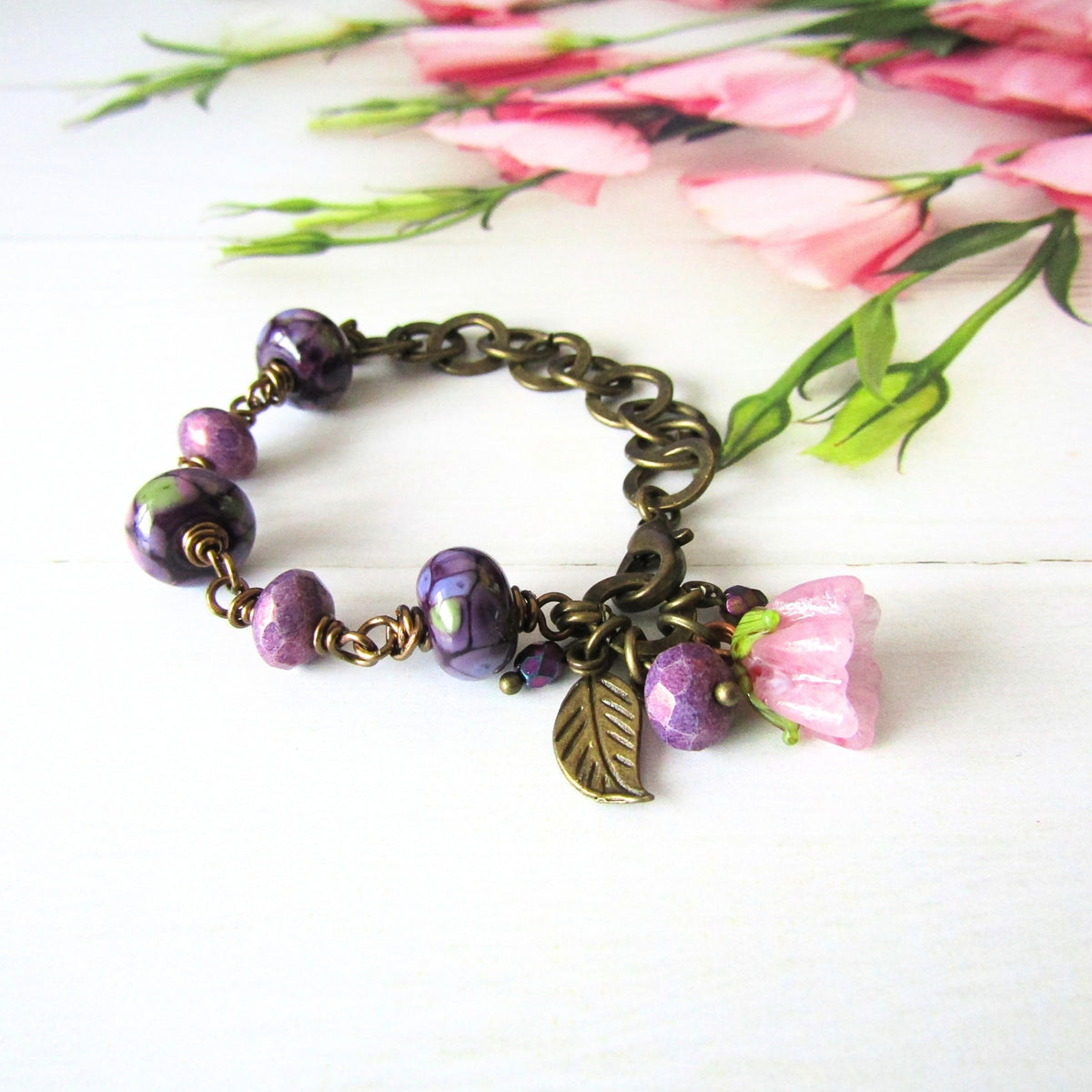 A handmade charm bracelet with purple glass beads, a pink flower charm, and a leaf charm, set against a white wooden background with pink flowers in the upper right.
