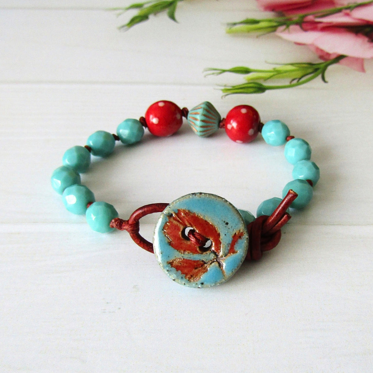 A handmade beaded bracelet with turquoise and red dotty beads, featuring a large blue & red button clasp with a leaf imprint design, displayed on a white wooden surface with pink flowers in the background.