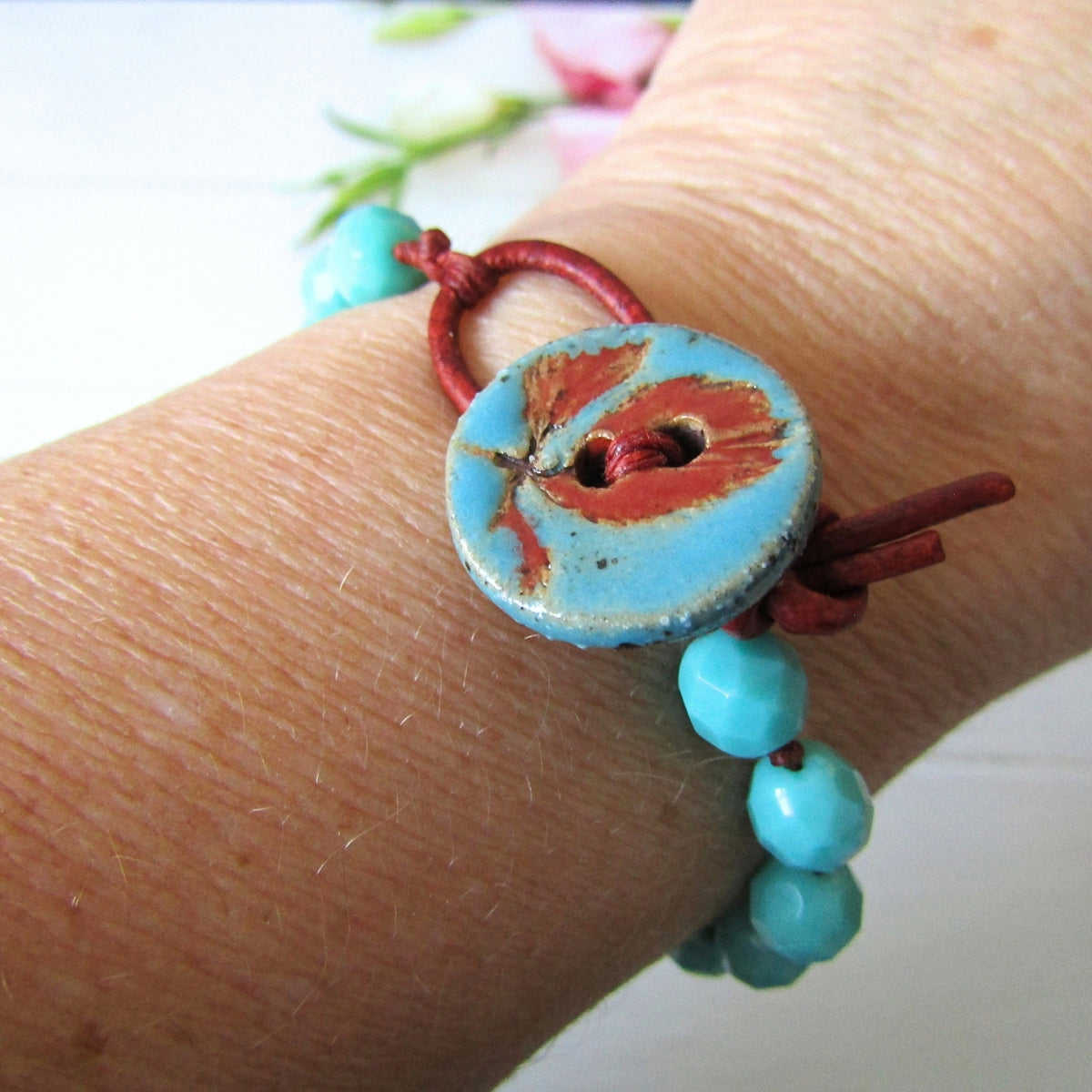 A close-up of a person's wrist wearing a handmade bracelet featuring a large turquoise and rust-red ceramic button, small turquoise beads, and a red cord wrap.