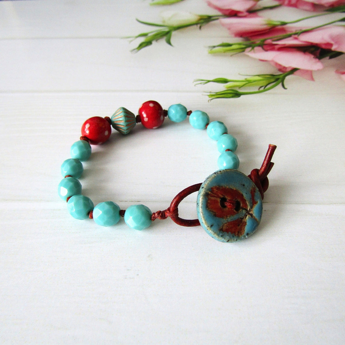 A handmade beaded bracelet with turquoise and red beads, featuring a large blue & red button clasp, displayed on a white wooden surface with pink flowers in the background.
