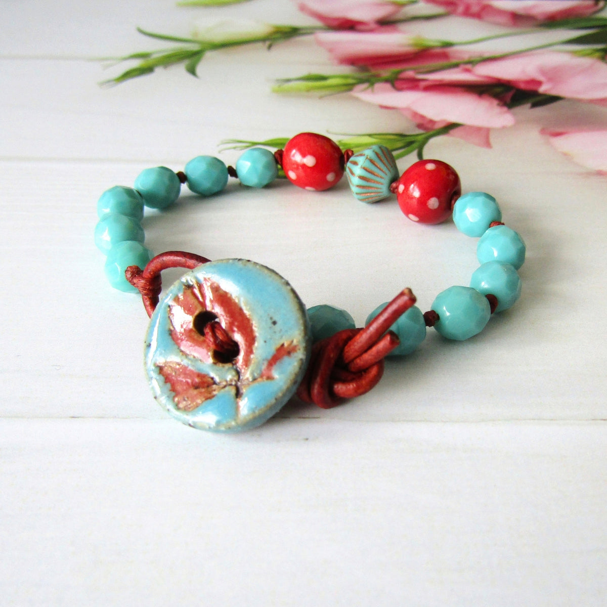 A handmade beaded bracelet with turquoise blue beads, red polka-dot beads, a patterned bicone bead, and a light blue & red leaf pattern button, tied with red cord on a white wooden surface with pink flowers in the background.