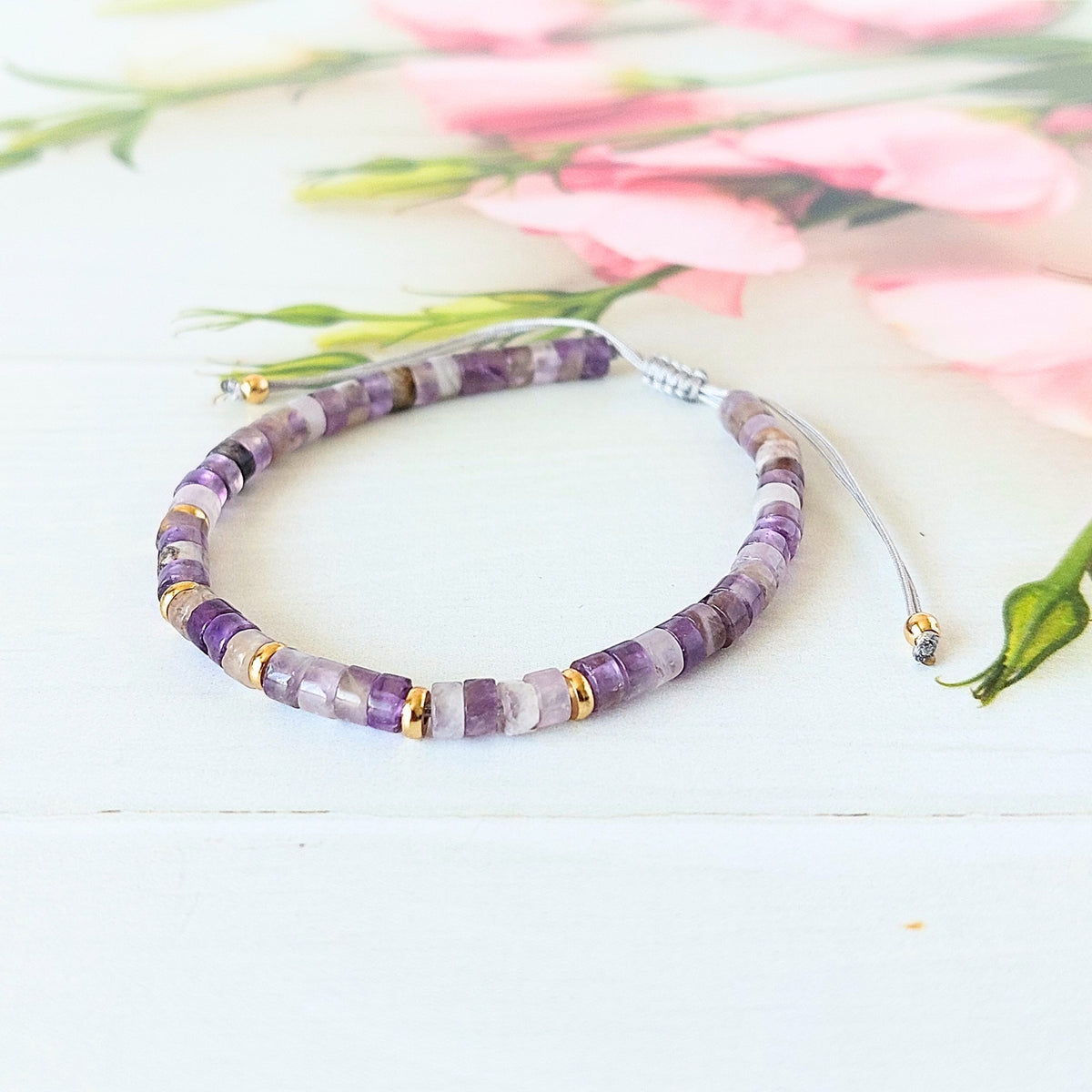 A beaded cord bracelet made of purple amethyst disc beads with small gold spacer rings, displayed on a white wooden surface with soft pink flowers in the background.