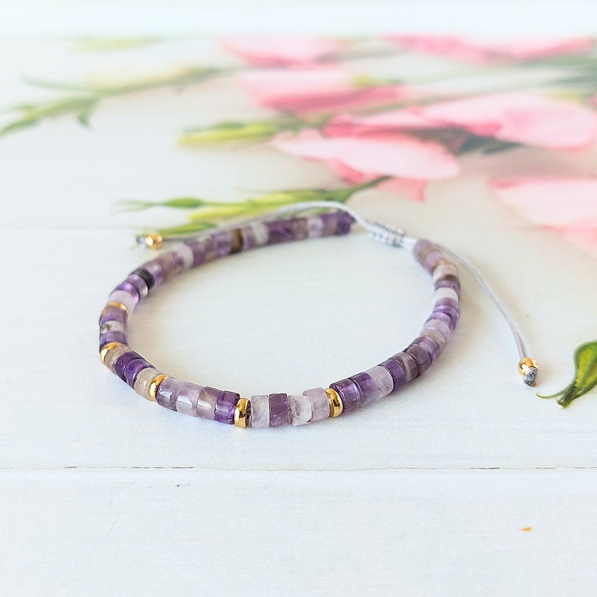 An amethyst beaded bracelet made of purple and white gemstone beads with small gold spacer accents, displayed on a white wooden surface with soft pink flowers blurred in the background.