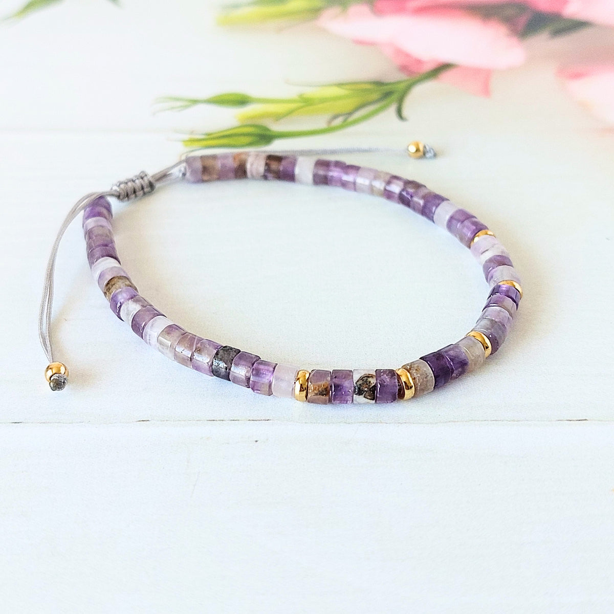 A beaded gemstone bracelet made of purple, white, and grey translucent beads with small gold spacer accents, displayed on a white wooden surface with soft pink flowers in the background.