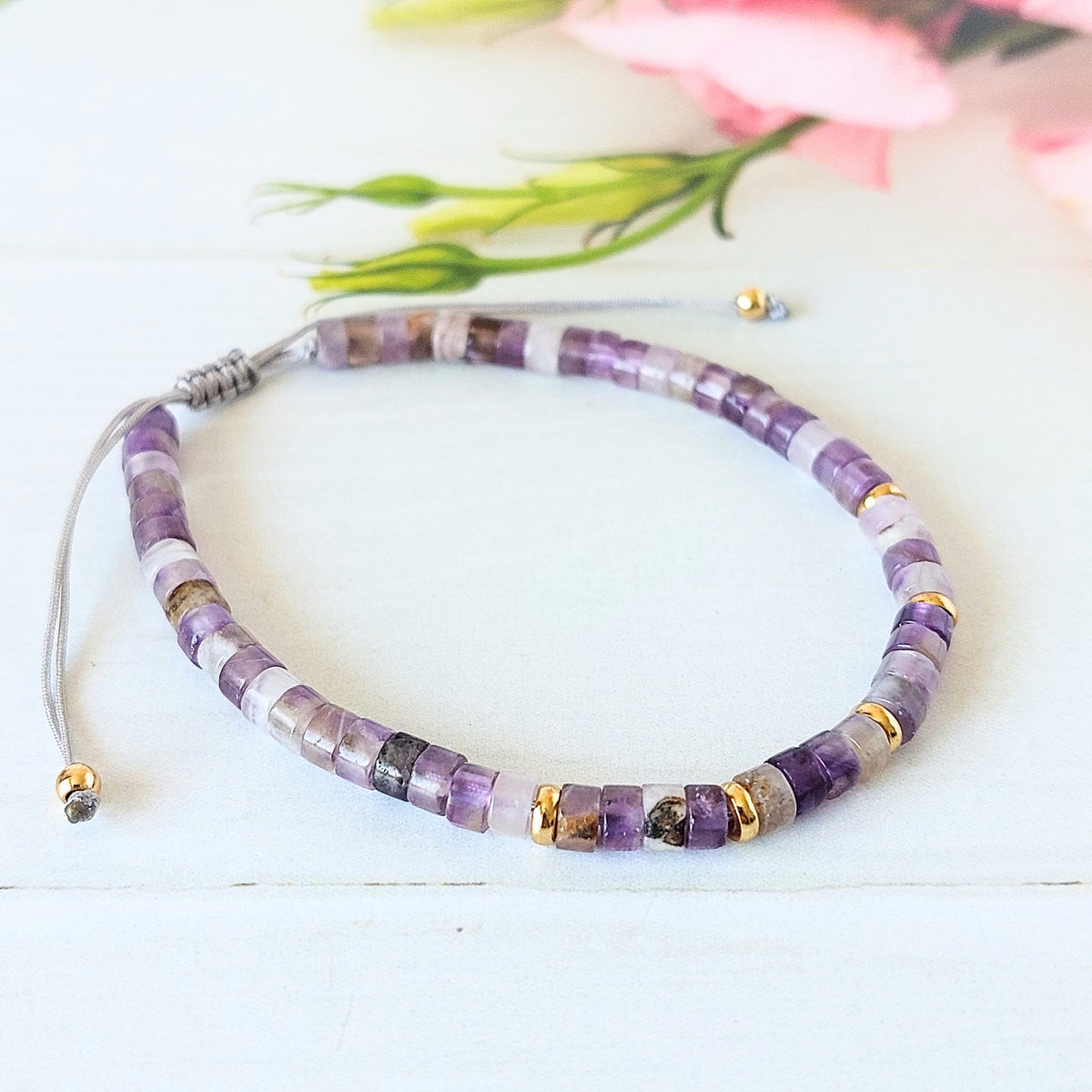 A beaded bracelet made of purple, white, and grey translucent beads with small gold spacer accents, displayed on a white wooden surface with soft pink flowers in the background.