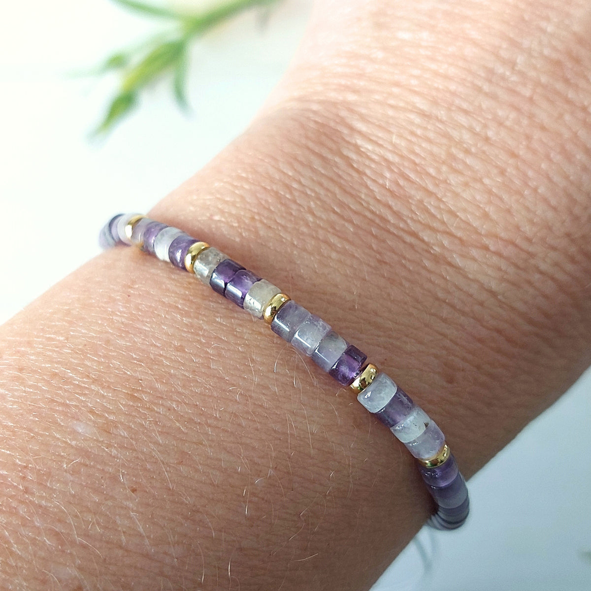 A close-up of a beaded bracelet worn on a wrist. The bracelet features alternating translucent purple and white amethyst beads separated by small gold spacer rings, set against a soft, out-of-focus light background.