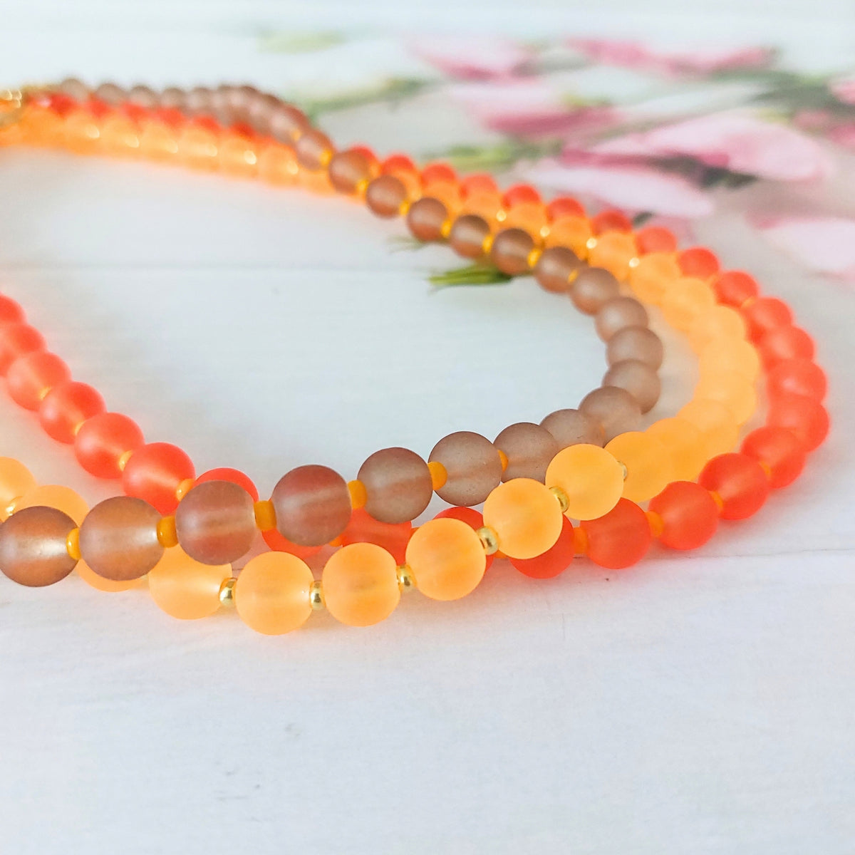 Three round beaded necklaces arranged in a circle on a white wooden surface. The necklaces feature warm brown beads, soft amber glass beads and lively coral-tone beads.