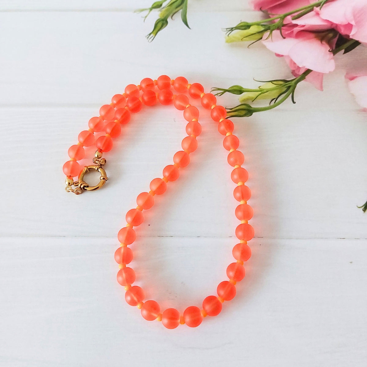 A beaded necklace made of frosted coral orange glass beads separated by small light orange spacer beads, with a gold clasp, arranged on a white wooden surface next to pink flowers.