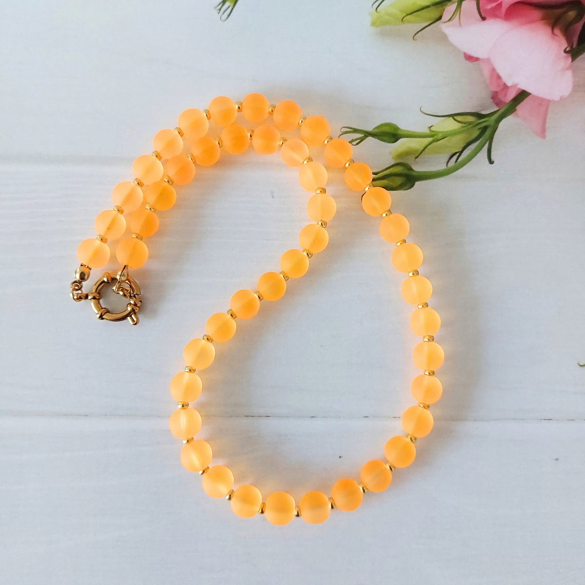An orange beaded necklace with a gold clasp, arranged on a white wooden surface next to pink flowers.